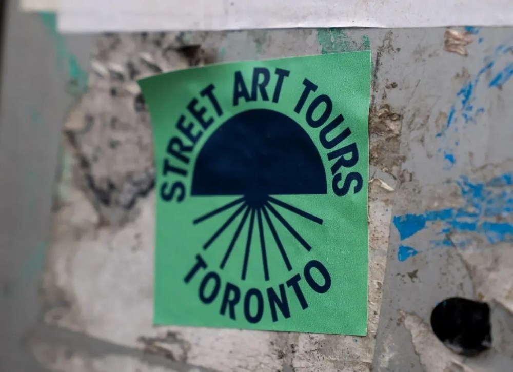 Green sticker on wall reading 'Street Art Tours Toronto' in blue letters with a sunburst design.