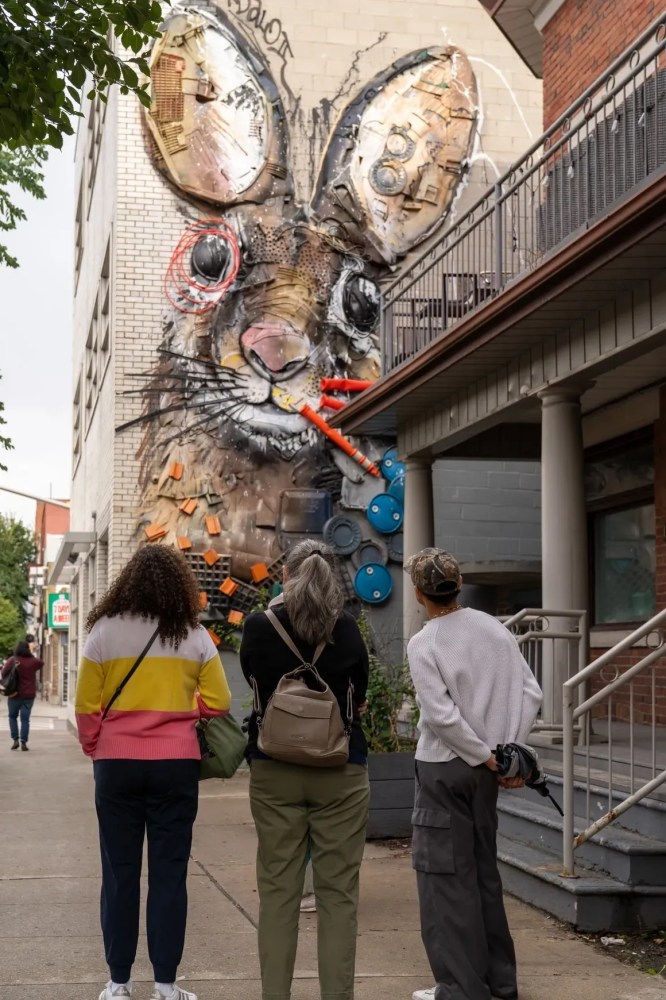 Three people view a large outdoor wall mural of a rabbit made from recycled materials.