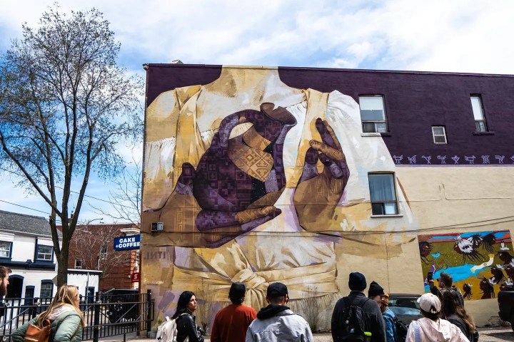 People viewing a large mural on a building under a cloudy sky.