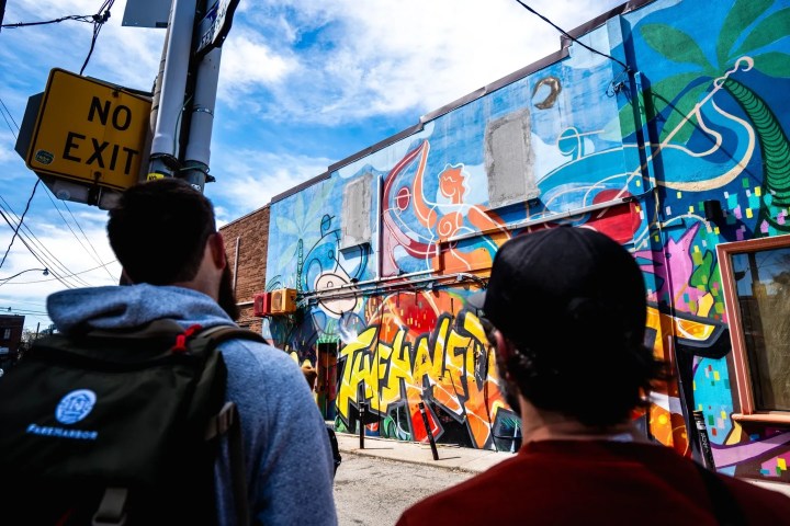 Two people view colorful graffiti mural in alley.